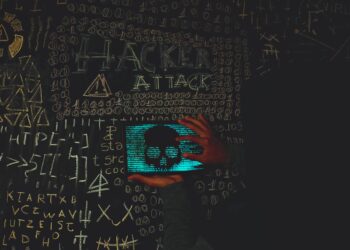 A hacker in a dark hoodie working on a computer with cryptocurrency symbols in the background.