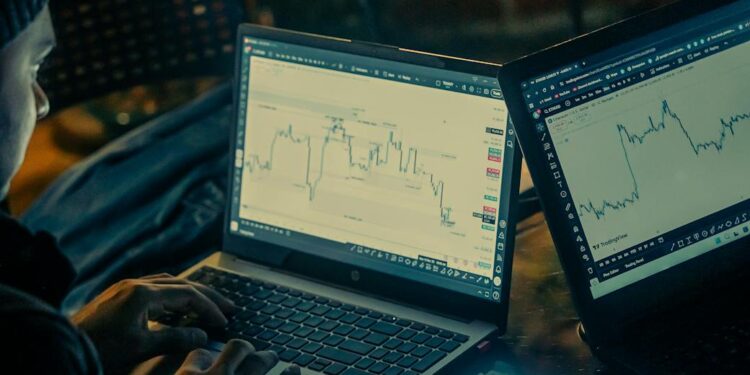 A trader monitors fluctuating cryptocurrency charts on multiple screens in a dimly lit room.