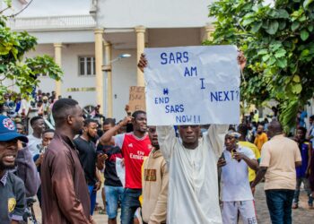 A user interacting with the Bitchat app on a smartphone amidst protest signs and unrest.