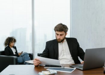 Retired man looking distressed while reviewing financial documents and a laptop with crypto data.