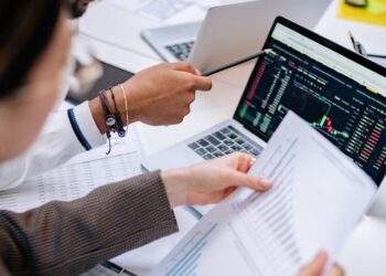 Two professionals discussing a document with cryptocurrency charts and financial data on a screen.