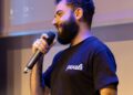 Joe Lubin speaking at a crypto conference, with visuals of DeFi logos in the background.