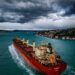 A cargo ship navigates the Strait of Hormuz with digital currency icons overlayed.