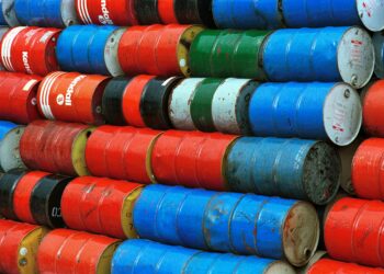 Oil barrels stacked at a port with a ship entering the Strait of Hormuz in the background.