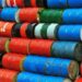 Oil barrels stacked at a port with a ship entering the Strait of Hormuz in the background.