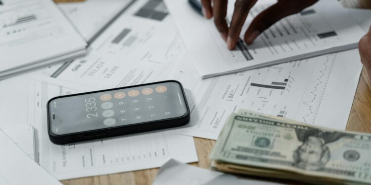 A hand holds a smartphone displaying a cryptocurrency graph and dollar bills on a table.