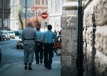 A handcuffed man being escorted by authorities outside a courthouse with Bitcoin symbols in the background.