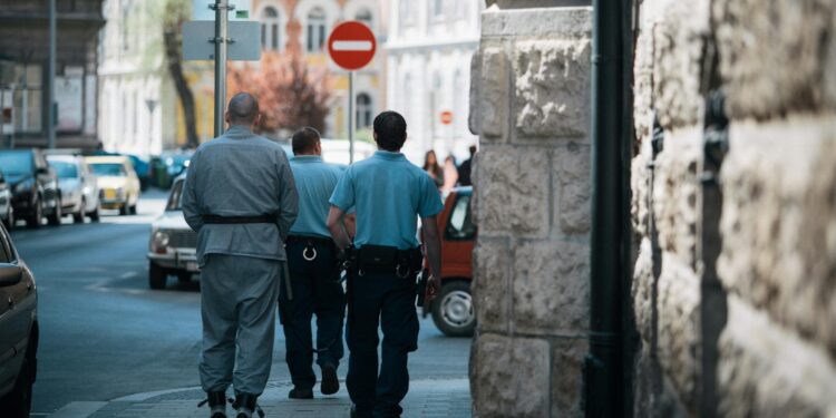A handcuffed man being escorted by authorities outside a courthouse with Bitcoin symbols in the background.