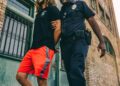 Young man in handcuffs being escorted by police in front of a crowd, reflecting crypto controversy.