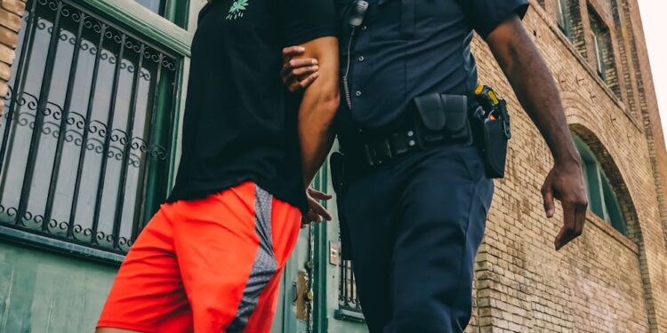 Young man in handcuffs being escorted by police in front of a crowd, reflecting crypto controversy.