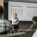 Sam Altman speaking at a conference, with graphs and charts on AI investments in the background.
